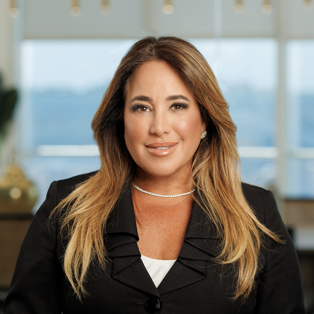 A woman with long light brown hair, wearing a black blazer over a white top and a pearl necklace, smiles at the camera in a modern office setting with blurred windows in the background. South Florida Business & Wealth