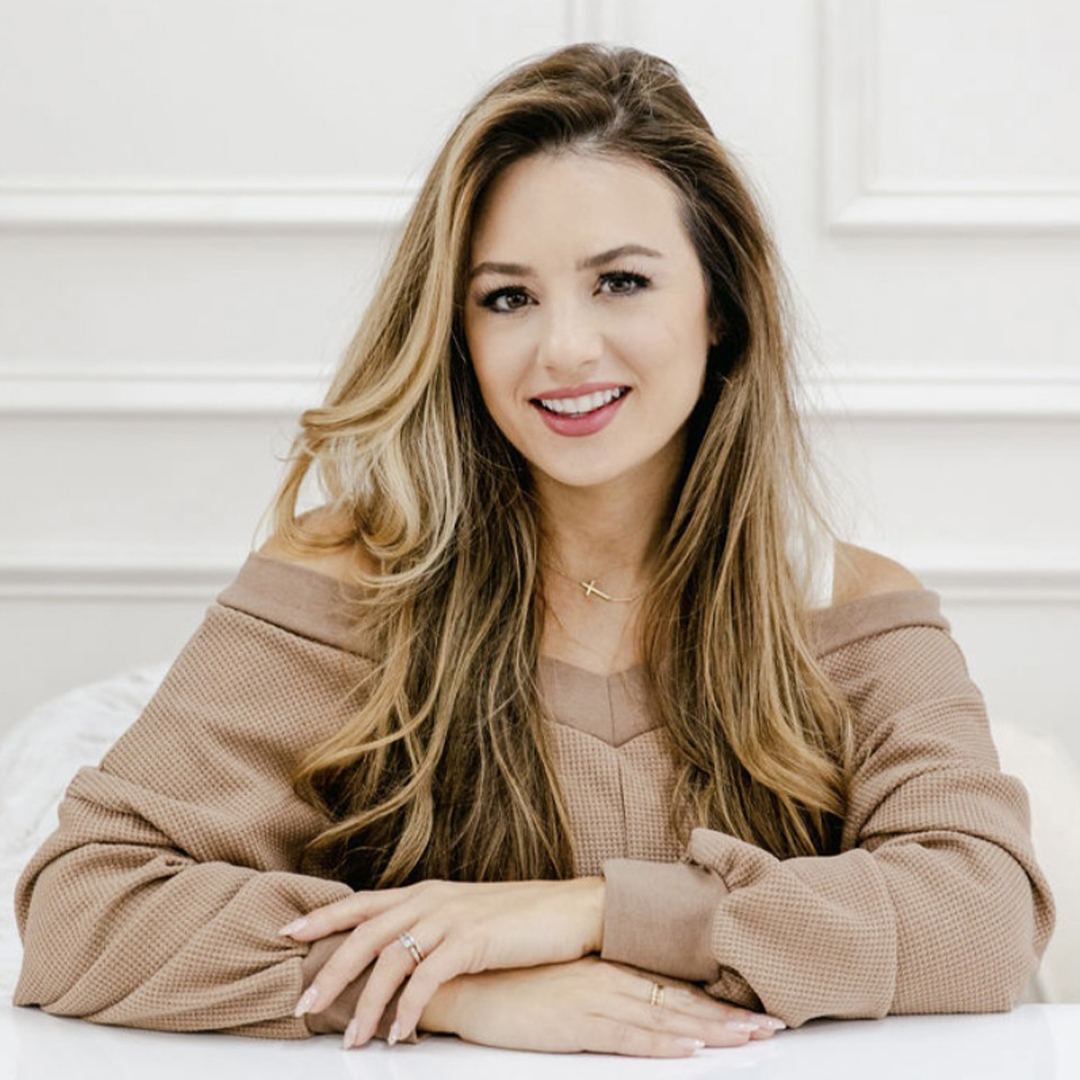 A woman with long, wavy brown hair sits at a table, smiling. She wears an off-the-shoulder tan sweater and a delicate necklace in a light, elegant room with white walls. South Florida Business & Wealth