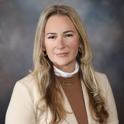 A woman with long blonde hair wearing a beige blazer, brown top, and white turtleneck, poses in front of a blurred gray and blue background. She has gold hoop earrings and a gold necklace, and is smiling slightly. South Florida Business & Wealth