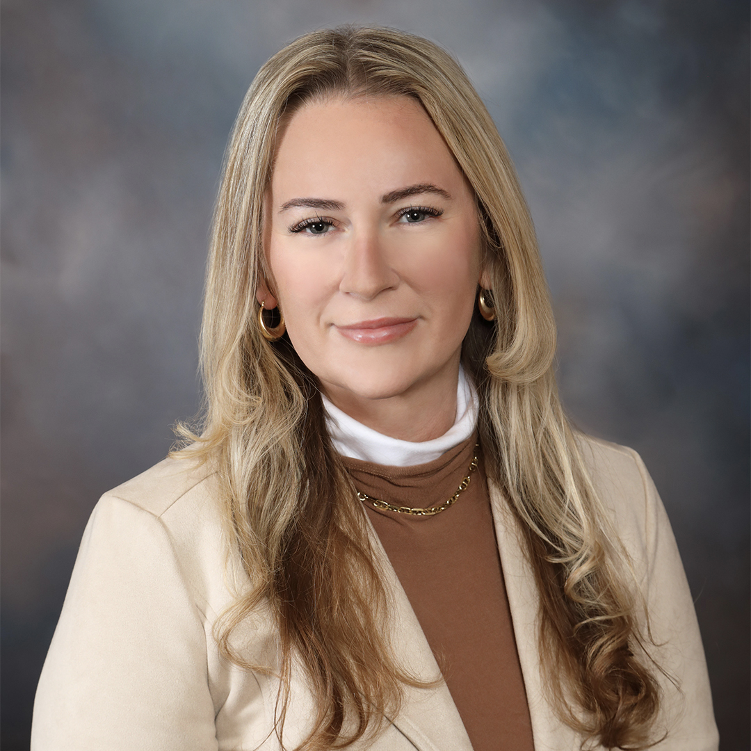 A woman with long blonde hair wearing a beige blazer, brown top, and white turtleneck, poses in front of a blurred gray and blue background. She has gold hoop earrings and a gold necklace, and is smiling slightly. South Florida Business & Wealth