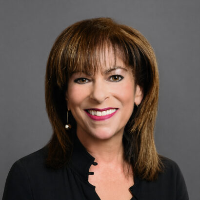 A woman with straight brown hair and bangs smiles at the camera. She is wearing a black top and a single dangling earring, posed against a plain gray background. South Florida Business & Wealth