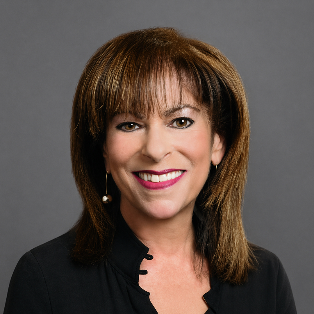 A woman with straight brown hair and bangs smiles at the camera. She is wearing a black top and a single dangling earring, posed against a plain gray background. South Florida Business & Wealth