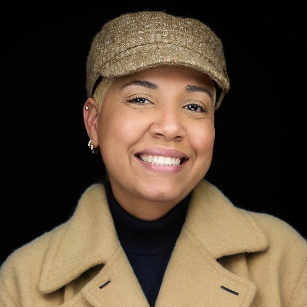 A smiling person wearing a tan wool coat and a matching textured cap, with a black turtleneck underneath, set against a plain black background. South Florida Business & Wealth
