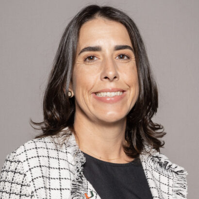 A woman with shoulder-length dark brown hair smiles at the camera. She is wearing a black top and a white checkered blazer, standing against a plain light gray background. South Florida Business & Wealth
