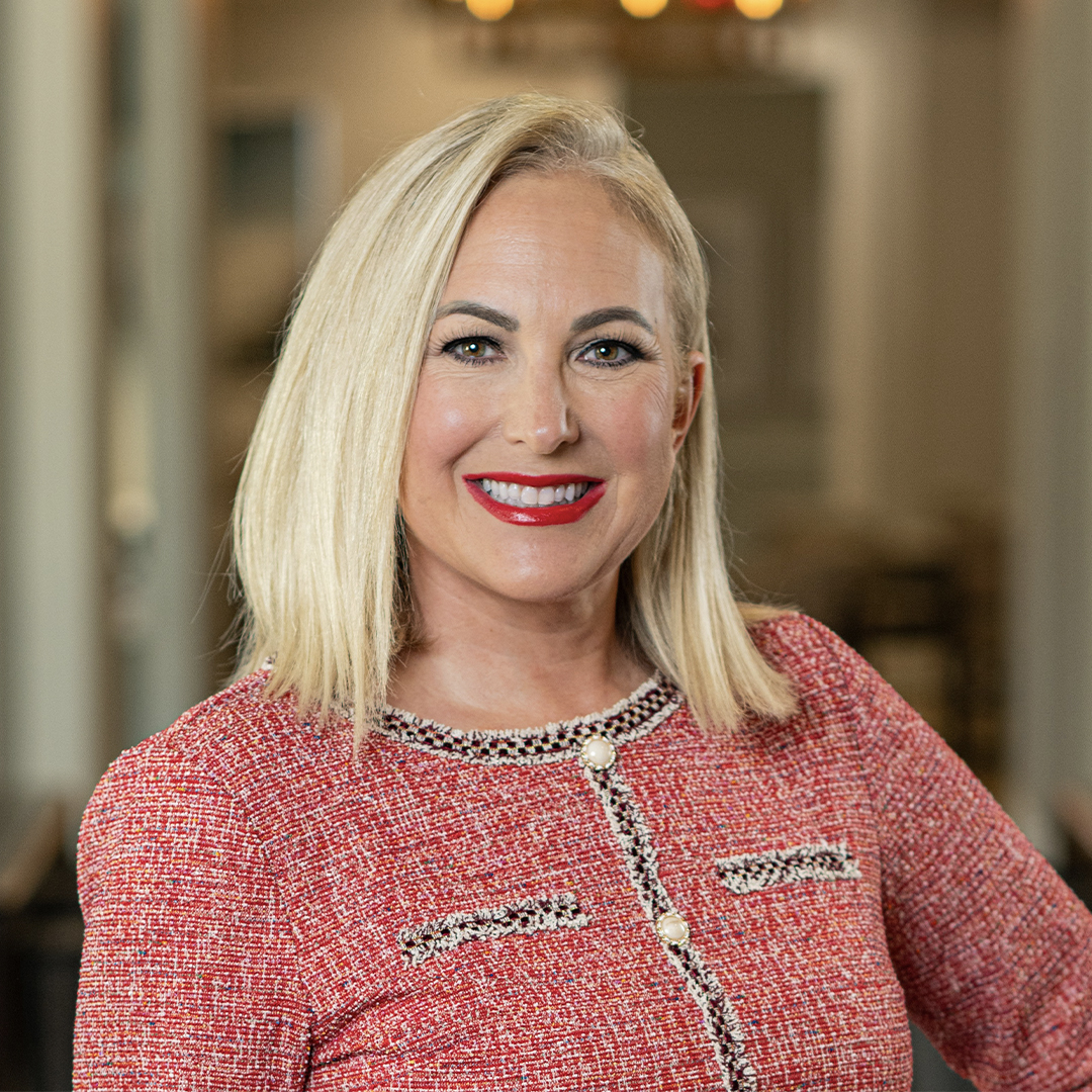 A woman with straight blonde hair wearing a textured red jacket with decorative trim, smiling and standing indoors, with a softly blurred background. South Florida Business & Wealth