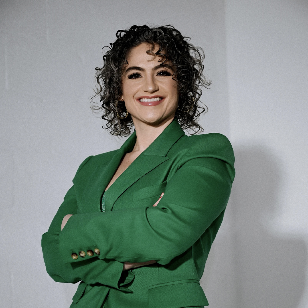 A smiling woman with curly dark hair wearing a green blazer stands with arms crossed against a plain light background. South Florida Business & Wealth