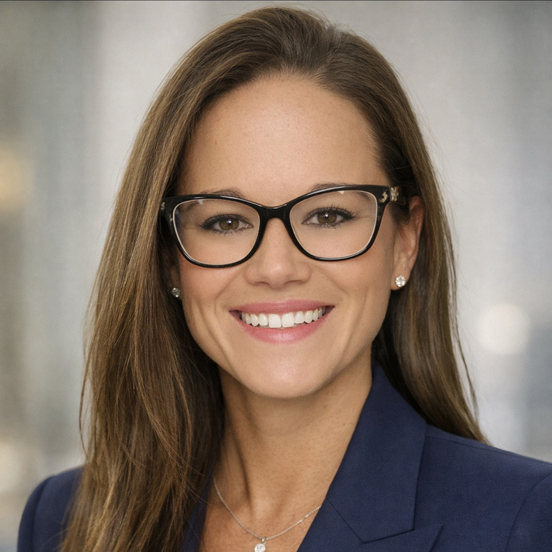 Woman with long brown hair and glasses smiling, wearing a navy blazer, pearl earrings, and a necklace. The background is softly blurred and neutral, suggesting a professional headshot. South Florida Business & Wealth