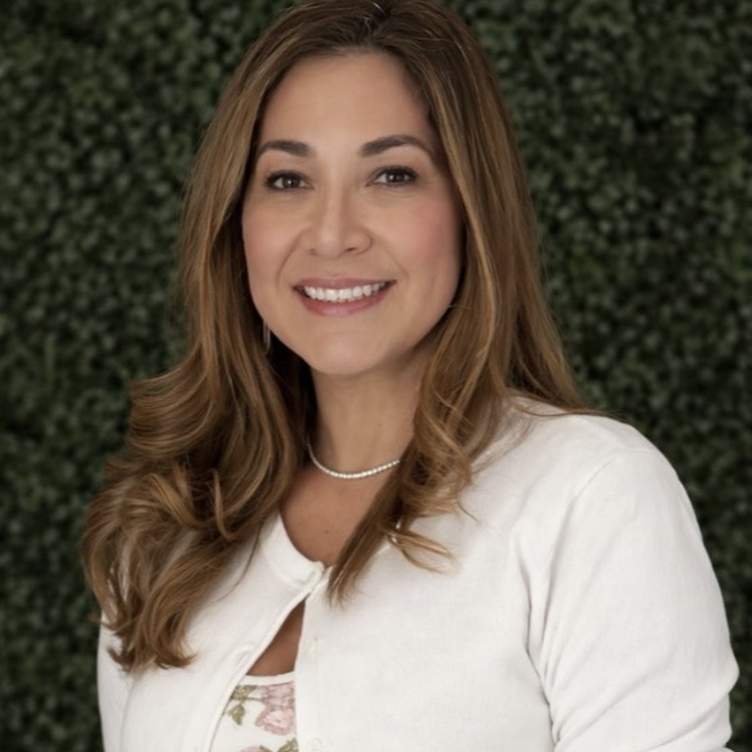 A woman with long, wavy light brown hair smiles at the camera. She is wearing a white top and a pearl necklace, standing in front of a green leafy background. South Florida Business & Wealth