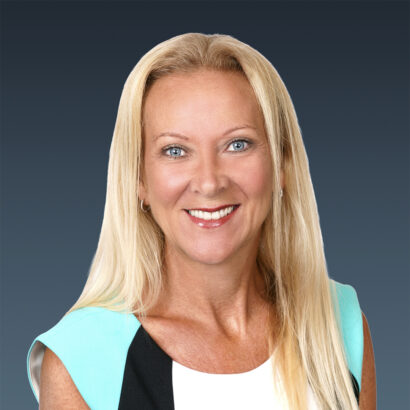 A woman with long blonde hair and blue eyes smiles at the camera. She is wearing a white, black, and turquoise top and is posed against a plain dark blue background. South Florida Business & Wealth