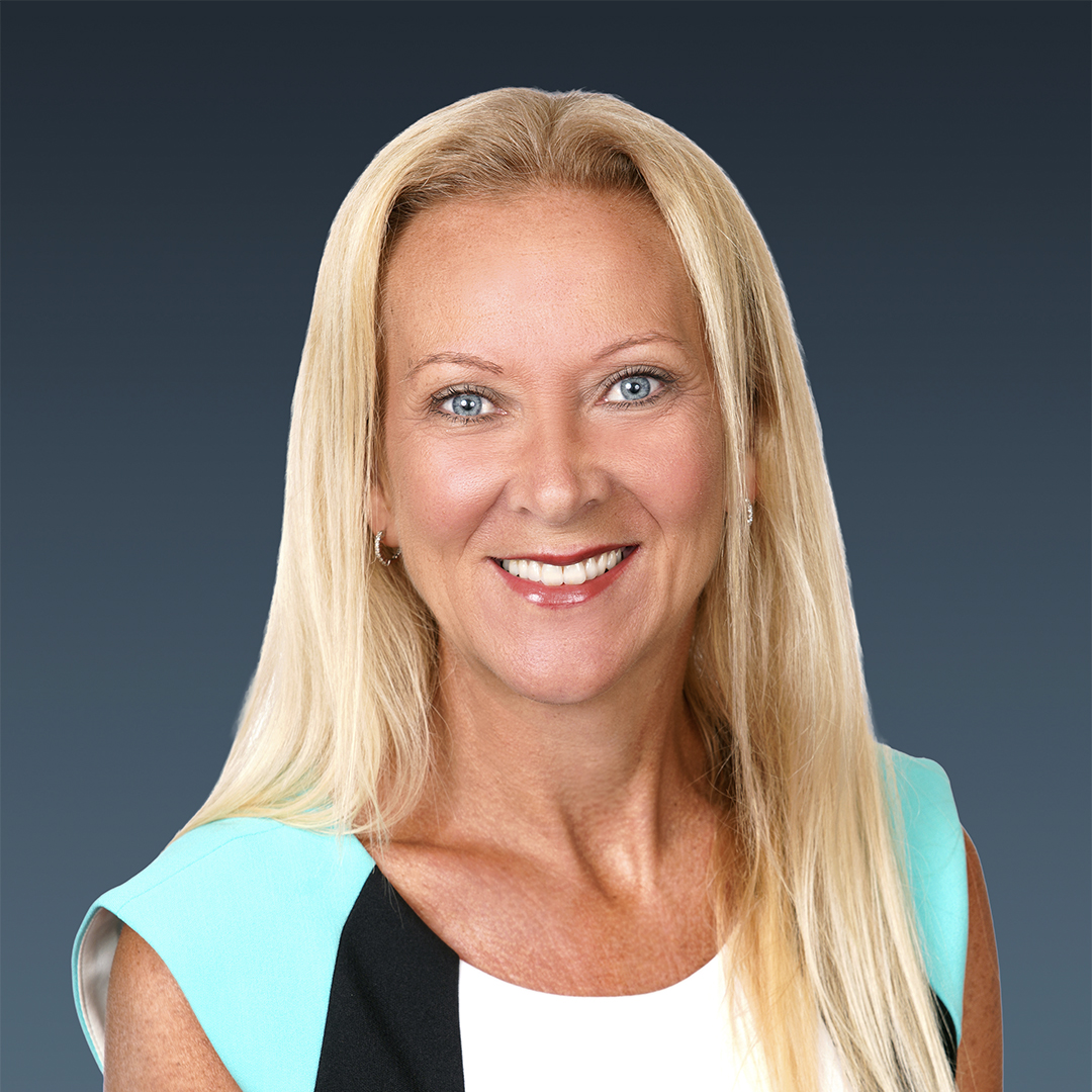 A woman with long blonde hair and blue eyes smiles at the camera. She is wearing a white, black, and turquoise top and is posed against a plain dark blue background. South Florida Business & Wealth