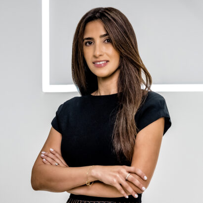 A woman with long brown hair wearing a black short-sleeve top stands with arms crossed, smiling slightly, in front of a gray background with a bright white geometric line. South Florida Business & Wealth