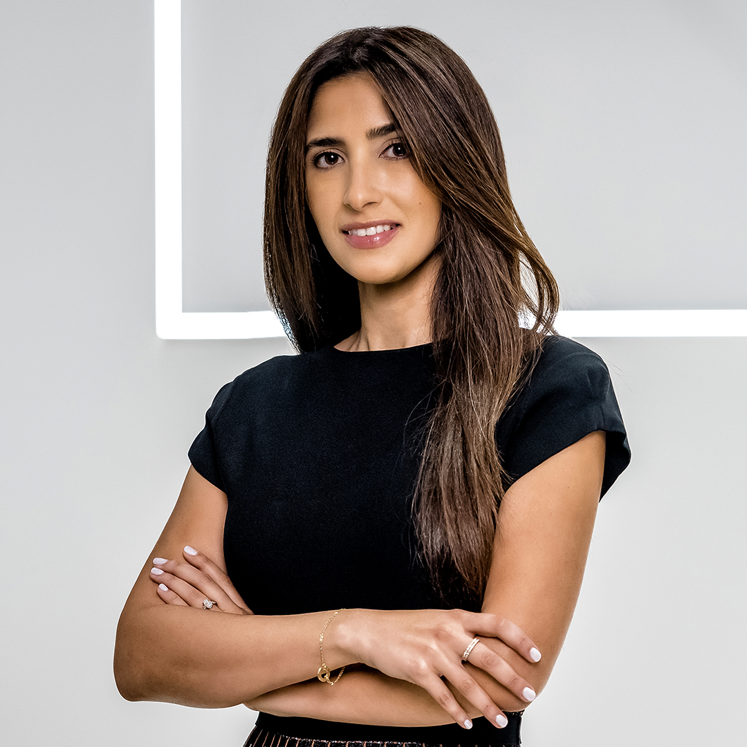 A woman with long brown hair wearing a black short-sleeve top stands with arms crossed, smiling slightly, in front of a gray background with a bright white geometric line. South Florida Business & Wealth