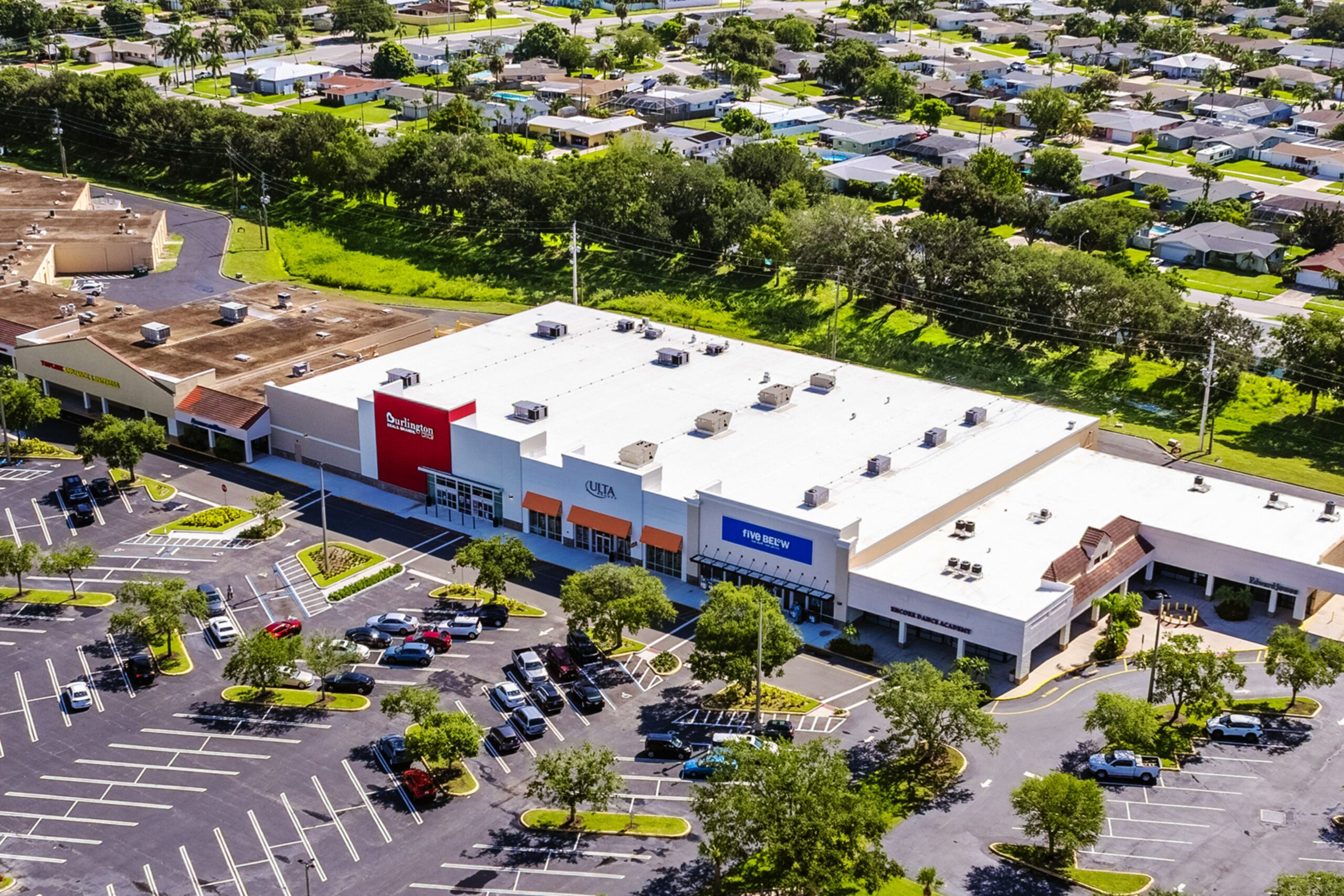 Aerial view of a shopping center with stores, including Ross Dress for Less, Ulta Beauty, and Five Below, in front of a large parking lot with scattered cars and a residential neighborhood in the background. South Florida Business & Wealth