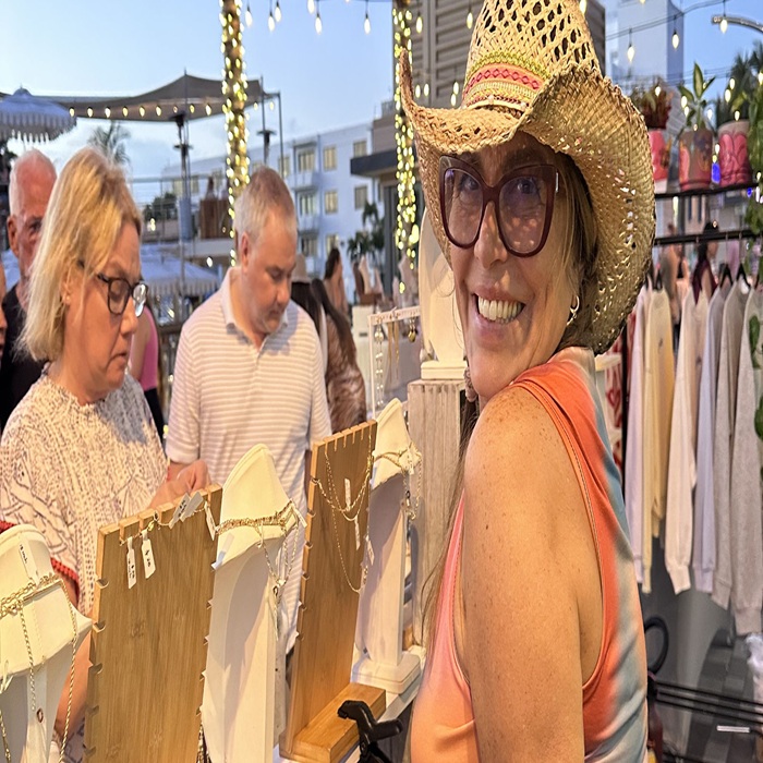 A smiling woman in a straw hat and sunglasses browses jewelry at an outdoor market stall, with other shoppers and string lights in the background. South Florida Business & Wealth
