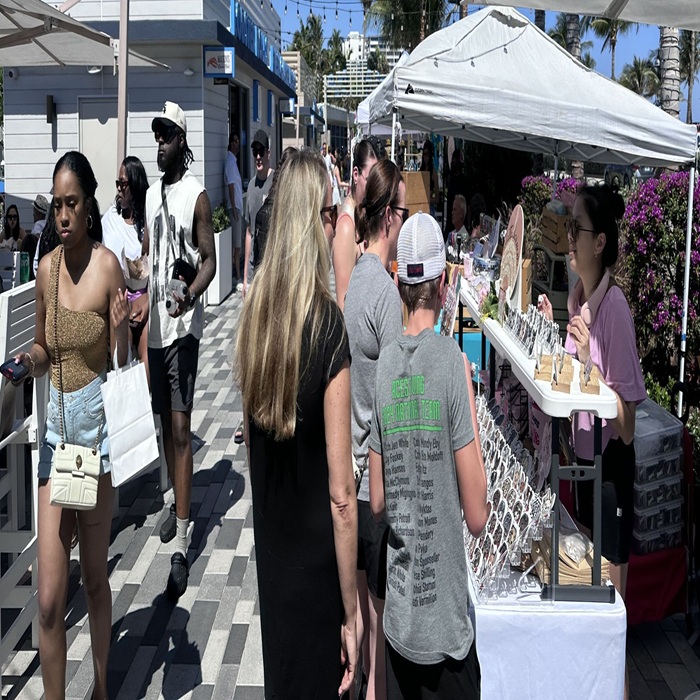People browse outdoor market stalls on a sunny day. Vendors display jewelry and accessories. Some shoppers hold drinks or bags, and palm trees are visible in the background. The atmosphere is busy and lively. South Florida Business & Wealth