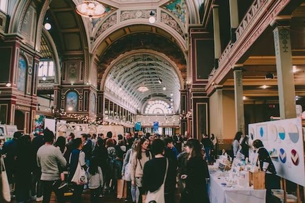 A large crowd browses booths and displays at an indoor market or fair inside an ornate, high-ceilinged hall with arches, decorative molding, and stained glass windows. South Florida Business & Wealth