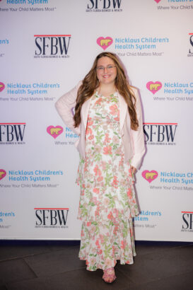 A woman with long brown hair and glasses smiles while posing in front of a step-and-repeat banner. She wears a floral dress, pale pink jacket, and matching shoes. Banners display Nicklaus Children’s Health System and SFBW logos. South Florida Business & Wealth