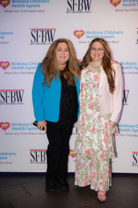 Two women stand smiling in front of a backdrop featuring Nicklaus Children’s Health System and SFBW logos. One wears a blue blazer and black pants; the other wears a pink floral dress with a white blazer. South Florida Business & Wealth