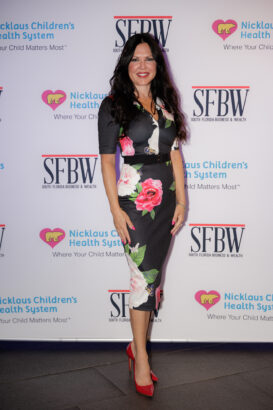 A woman with long dark hair in a floral dress and red heels poses in front of a backdrop with the logos for Nicklaus Children’s Health System and SFBW (South Florida Business & Wealth). South Florida Business & Wealth