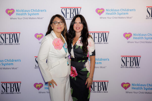 Two women stand together smiling in front of a step-and-repeat backdrop featuring logos for Nicklaus Children's Health System and SFBW. One woman wears a white suit with floral details, and the other wears a black floral dress. South Florida Business & Wealth