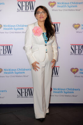 A woman in a white suit with a large pink flower pin, patterned blue top, and glasses stands on a gray carpet in front of a step-and-repeat banner for SFBW and Nicklaus Children's Health System. South Florida Business & Wealth