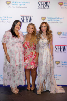 Three women wearing floral dresses smile and pose together in front of a step-and-repeat banner featuring the logos of Nicklaus Children’s Health System and SFBW (South Florida Business & Wealth). South Florida Business & Wealth