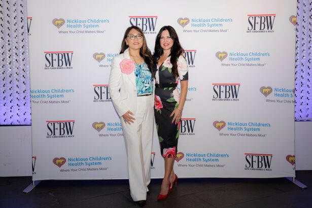 Two women posing and smiling on a red carpet in front of a step-and-repeat backdrop with logos for SFBW and Nicklaus Children's Health System. Both are dressed in floral outfits. South Florida Business & Wealth