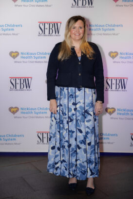 A woman with long blonde hair, wearing a navy cardigan over a blue and white floral dress, stands smiling in front of a step-and-repeat banner for Nicklaus Children's Health System and SFBW. South Florida Business & Wealth