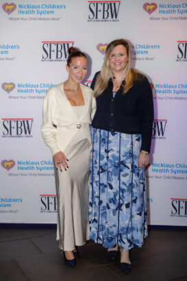 Two women stand smiling in front of a backdrop displaying the logos of Nicklaus Children’s Health System and SFBW. One wears a cream sweater over a silk dress; the other wears a navy blazer and blue floral skirt. South Florida Business & Wealth