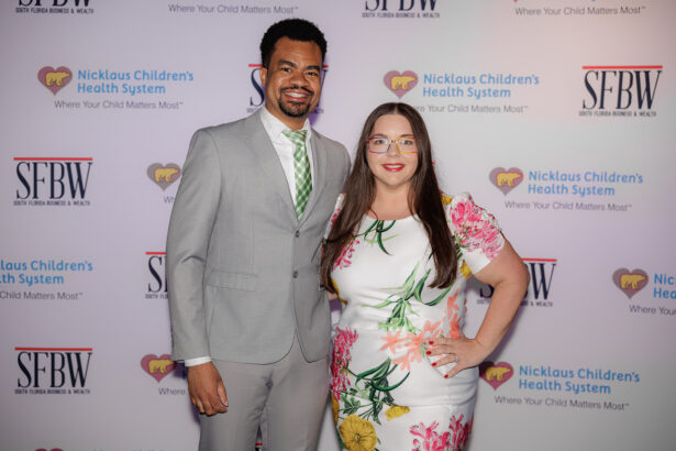 A man in a light gray suit and green tie stands next to a woman in a white floral dress. They are smiling and posing in front of a step-and-repeat banner with Nicklaus Children's Health System and SFBW logos. South Florida Business & Wealth