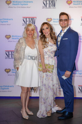 Three people pose together at an event in front of a step-and-repeat banner. Two women wear elegant white dresses, one with floral patterns; a man wears a blue suit and glasses. All are smiling. South Florida Business & Wealth