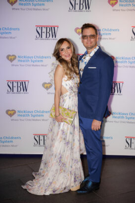 A woman in a floral dress and a man in a blue suit pose together, smiling, on a step-and-repeat backdrop featuring logos for Nicklaus Children's Health System and SFBW. South Florida Business & Wealth