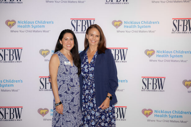 Two women smiling and standing together in front of a step-and-repeat backdrop with logos for Nicklaus Children’s Health System and SFBW. Both women are wearing blue patterned dresses, and one has a blue blazer. South Florida Business & Wealth