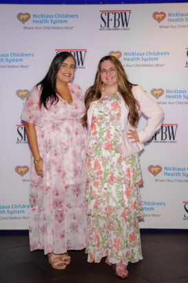 Two women pose together, both wearing long floral dresses and light-colored jackets. They stand in front of a backdrop featuring logos for Nicklaus Children’s Health System and SFBW. South Florida Business & Wealth