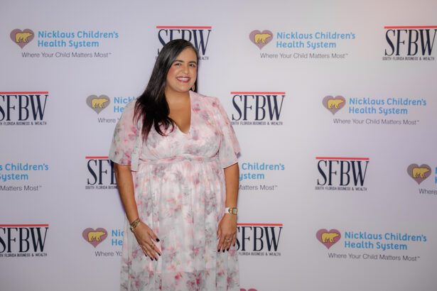 A woman in a pink floral dress smiles while posing in front of a step-and-repeat backdrop with logos for Nicklaus Children's Health System and SFBW magazine. South Florida Business & Wealth