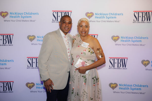 A man in a light suit and a woman in a light floral dress smile together on a step-and-repeat backdrop displaying Nicklaus Children’s Health System and SFBW logos. South Florida Business & Wealth