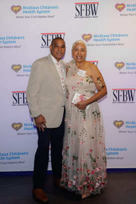 A man in a light suit and a woman in a floral, sleeveless dress pose together, smiling, in front of a step-and-repeat banner for Nicklaus Children’s Health System and South Florida Business & Wealth. South Florida Business & Wealth