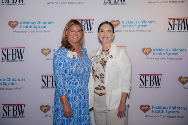 Two women stand smiling in front of a backdrop featuring the Nicklaus Children’s Health System and SFBW logos at an event. One wears a blue dress, the other a white blazer over a patterned shirt. South Florida Business & Wealth