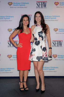 Two women posing and smiling at an event, one wearing a red dress and black heels, the other wearing a white floral dress and beige heels, standing in front of a backdrop with “Nicklaus Children’s Health System” and “SFBW” logos. South Florida Business & Wealth