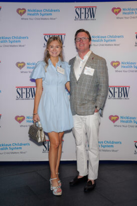 A woman in a light blue dress and a man in a plaid blazer and white pants pose together, smiling, in front of a step-and-repeat banner for Nicklaus Children’s Health System and SFBW. South Florida Business & Wealth