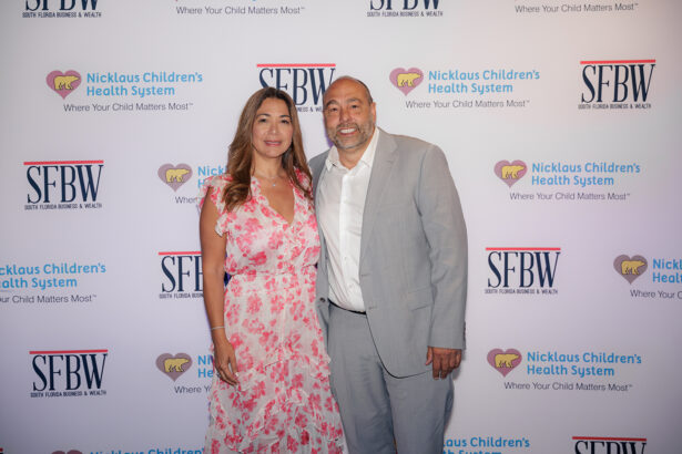 A woman in a pink floral dress and a man in a gray suit pose together, smiling, in front of a step-and-repeat backdrop with logos for Nicklaus Children’s Health System and South Florida Business & Wealth. South Florida Business & Wealth