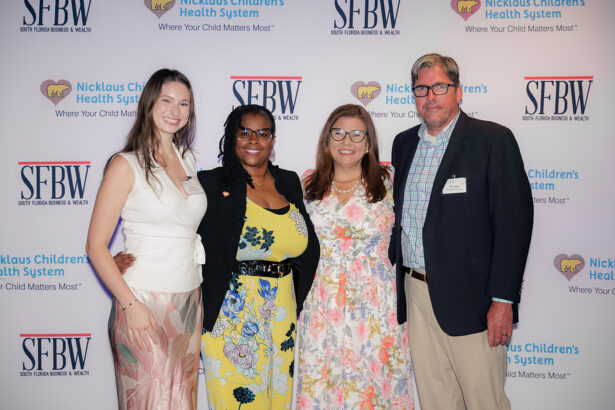 Four people stand smiling in front of a step-and-repeat banner with logos for Nicklaus Children's Health System and SFBW. Three women wear dresses and a man wears a blue blazer and khaki pants. South Florida Business & Wealth