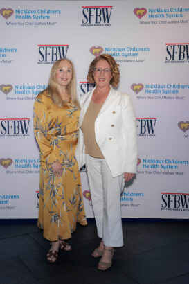 Two women pose and smile in front of a step-and-repeat banner for Nicklaus Children's Health System and SFBW. One wears a gold floral dress and heels, the other a white blazer with beige pants and sandals. South Florida Business & Wealth