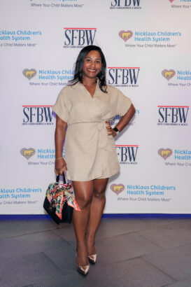 A woman in a beige dress and metallic heels poses, smiling, on a step-and-repeat backdrop with logos for Nicklaus Children’s Health System and SFBW. She holds a black handbag with a colorful scarf tied to it. South Florida Business & Wealth