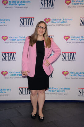 A woman with long blonde hair and glasses smiles while posing in a black dress and pink blazer. She stands in front of a step-and-repeat backdrop with logos for SFBW and Nicklaus Children’s Health System. South Florida Business & Wealth