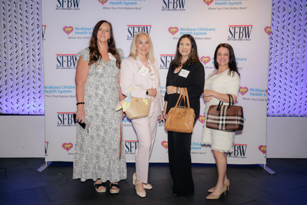 Four women dressed in business attire stand together, each holding handbags, posing for a photo in front of a backdrop featuring SFBW and Nicklaus Children’s Health System logos. South Florida Business & Wealth