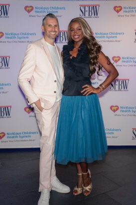 A man in a light pink suit and white sneakers stands beside a smiling woman in a black ruffled top and blue skirt. They pose in front of a step-and-repeat banner for Nicklaus Children's Health System and SFBW. South Florida Business & Wealth