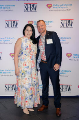 A woman in a floral dress and a man in a suit with a patterned shirt pose together, smiling, in front of a step-and-repeat backdrop with logos for SFBW and Nicklaus Children's Health System. South Florida Business & Wealth