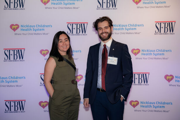 Two people standing and smiling in front of a step-and-repeat banner featuring logos for Nicklaus Children's Health System and SFBW at an event. The woman wears a sleeveless dress; the man wears a suit and tie. South Florida Business & Wealth
