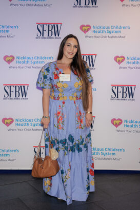 A woman in a long, blue floral dress stands smiling on a step-and-repeat backdrop featuring Nicklaus Children’s Health System and SFBW logos. She carries a tan handbag and has long brown hair. South Florida Business & Wealth